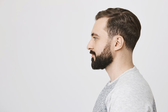 Profile portrait of handsome bearded adult european man with trendy haircut, standing over gray background. Hairdresser makes photo of new haircut he did for visitor to post it on site