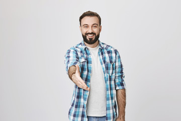Fototapeta premium Young cheerful bearded man with trendy haircut smiling broadly while stretching hand at camera in greeting gesture, standing over gray background. Nice to meet you, I am new in this company