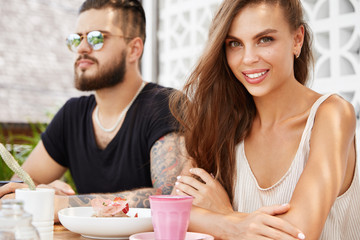 Obraz na płótnie Canvas Beautiful female with long hair and bearded stylish man in sunglasses sit together at cafe, eat dessert, like communication, travel together in exotic country, have much impressions, positive emotions