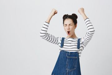 Brunette girl with hairbuns in striped top excited and glad to achieve victory, clenches fists,...