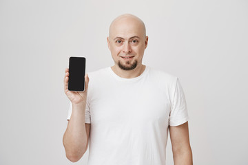 Indoor shot of handsome bald bearded guy holding black smartphone while smiling and looking pleased at camera, standing over gray background. Wealthy man bought his son phone for good marks at school