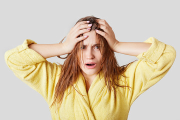Fototapeta premium Frustrated female touches head, has wet hair and disorder, doesn`t know what to wear and which hairstyle make for meeting, dressed in casual domestic clothing, isolated over white background