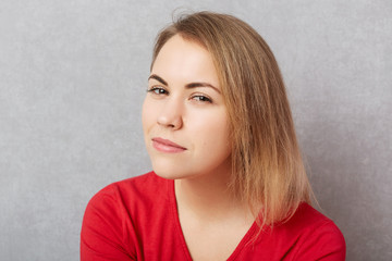 Horizontal shot of beautiful young blonde woman with intriguing cunning look, plans to do something unexpected, isolated over grey background. Pretty female poses indoor. Sly look at camera.