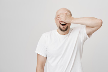 Astonished and excited bald bearded man with broad smile covering eyes with hand and awaiting for something positive to happen, standing over gray background. Birthday guy waits for his present