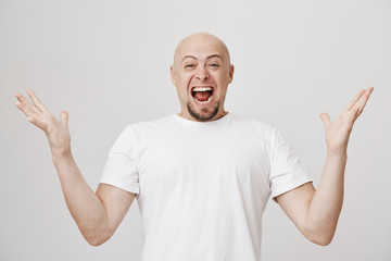 Naklejka premium Studio portrait of overwhelmed and excited bald caucasian man with beard shouting of happiness and spreading hands, showing his joy because of success over gray background. Guy became father