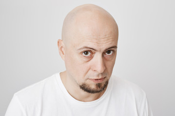 Close-up portrait of handsome bald man with fancy beard, staring up at camera with serious and confident look, standing over gray background. Tired office manager waits till his shift will over.