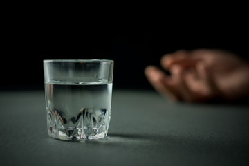 glass of vodka or alcohol drink, hand of a drunk man in the background, alcoholism and alcohol abuse concept, defocused, selective focus, close up, gray table, dark background