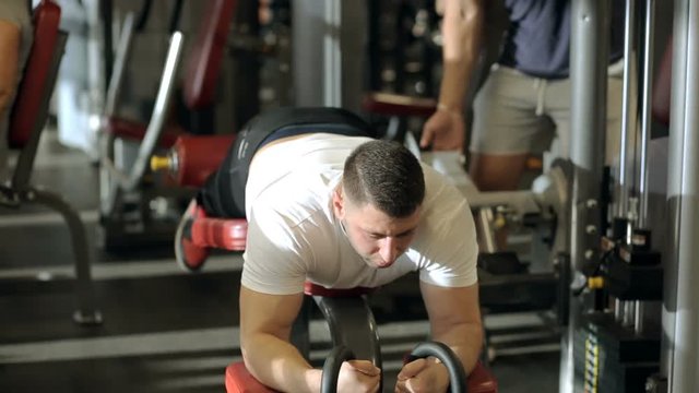 The athlete works on his hamstrings using exerciser in the modern gym. The sportsman lies on the red leather couch, holds the handles by the hands and curls his legs. The man wears white t-shirt and