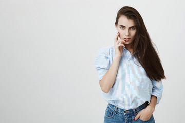 Hot and sensual european woman in blue-collar shirt flirting and glancing at camera with hand near face and another in pocket, standing over gray background. Famous actor poses for popular magazine