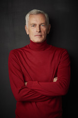 Casual senior man portrait. Handsome elderly man wearing red turtle neck sweater and looking at camera while standing against at dark background.