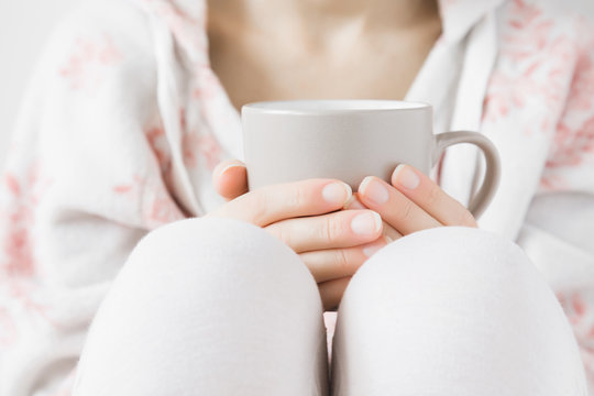 Woman in white soft home sweater holding a mug in hands at home. Enjoying coffee or tea in day off. Morning drink concept.