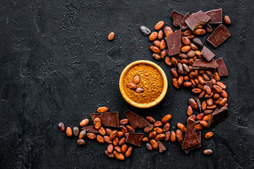 Chocolate and cacao concept. Cocoa powder in bowl near cocoa beans and broken chocolate on black background top view copy space