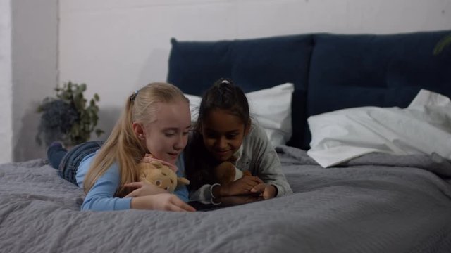 Cute multi ethnic little girls with plush toys working on digital tablet while lying on the bed in domestic room. Carefree multiracial kids surfing the net with touchpad and spending leisure at home.
