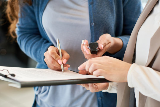 Close-up shot of unrecognizable customer holding car key in hand and signing contract after successful deal in automobile showroom, consultant assisting her
