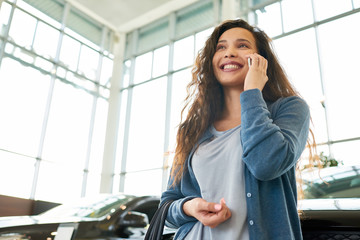 Low angle view of attractive mixed race woman looking away with charming smile and talking to her friend on smartphone while choosing new car in showroom, portrait shot