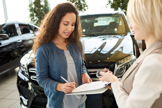 Portrait shot of pretty curly customer signing contract after successful deal in automobile showroom, blond-haired saleswoman passing car key to her