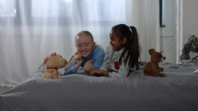Excited cute multi ethnic preteen girls having fun and playing on the bed at home. Carefree multiracial kids enjoying leisure together ,playing , tickling and laughing in domestic room.