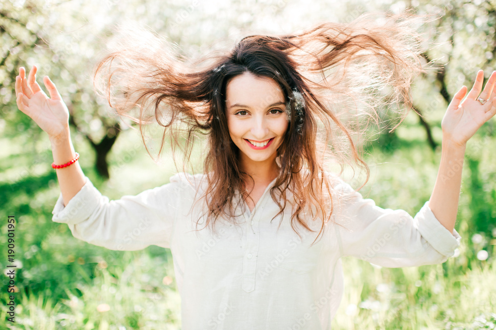 Wall mural Expressive portrait of young cheerful emotional model girl throwing hair. Spring is coming. Cute funny unusual female person enjoying innocence of life outdoor in blooming garden with blossom flowers.