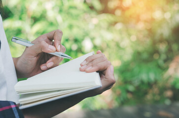 gorgeous woman hand writing on notebook paper with pen against green nature background and sun light.