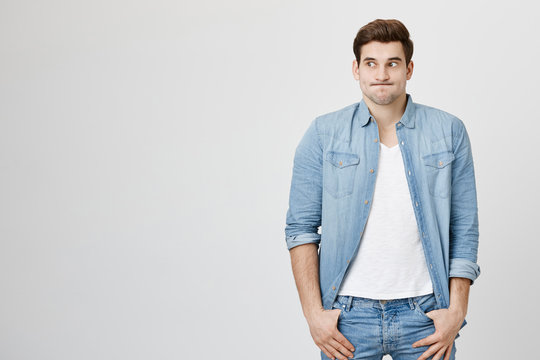 Studio shot of emotional clueless man with trendy haircut in denim shirt over white t-shirt having confused puzzled look, pouting cheeks, shrugging shoulders as he doesn't know reason of accident