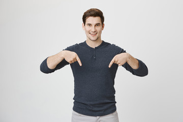 Attractive young boy with trendy haircut smiling and pointing down with both index fingers, standing over gray background. European model makes advertisement for business company