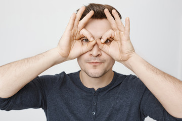 Close-up portrait of funny young guy making stupid face while showing glasses with hands and looking through it, standing over gray background. I am watching you. Man imitates he looks in binocular