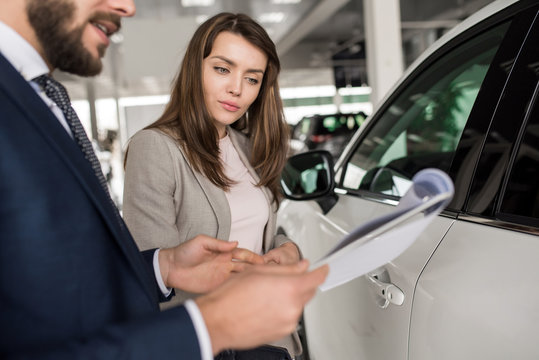 Portrait of modern young woman listening to car dealer while choosing luxury car in showroom