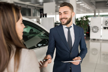 Portrait of handsome car salesman giving keys to young woman standing next to white shiny luxury car in dealership showroom
