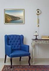 Vintage Furniture - Interior composition of retro blue armchair, vintage wooden beige table, table lamp, books, and pendulum clock over off white wall, tiled beige floor and orange ornate carpet