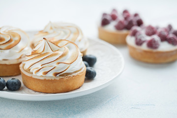 Delicious lemon and raspberry tartlets with meringue on a white vintage plate. Sweet treat on a light blue background. Flat lay and copy space. Top view