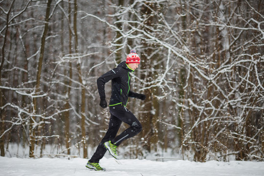 Image from side of man in sportswear, red cap on run in winter