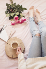 Young Caucasian woman in bed early in the morning, wearing distressed jeans and a white sweater and holds a cup of coffee. Roses and notebooks around. Cozy blogger mornings