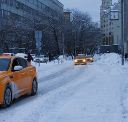 A yellow taxi rides along the winter road in the city after a snowfall. Twilight. A lot of snow. ...