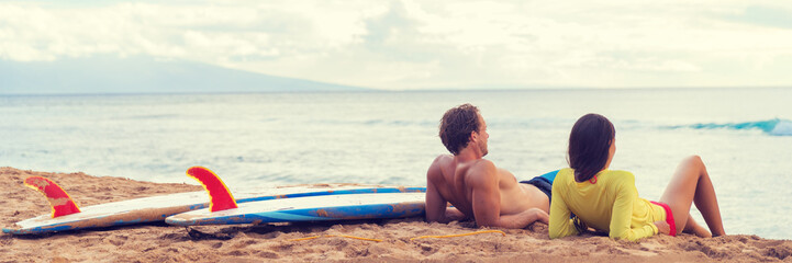 Couple surfers relaxing after surfing on hawaiian beach. Two people lying down on sand beach at sunset next to surfboards after a surf class in Hawaii, panorama banner crop.