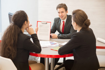 Group of business people sitting in circle at round meeting table and discussing work, focus on young businessman presenting contract to partners