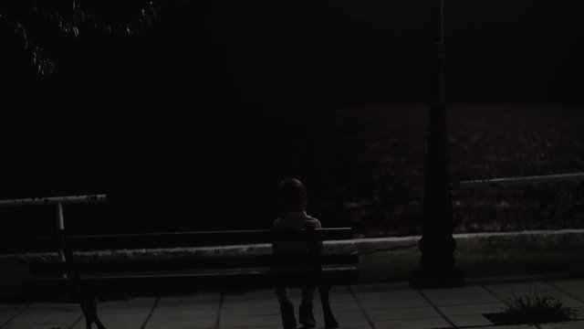 Bright lantern light in the darkness with following view to the lonely child sitting on the bench on waterfront at night