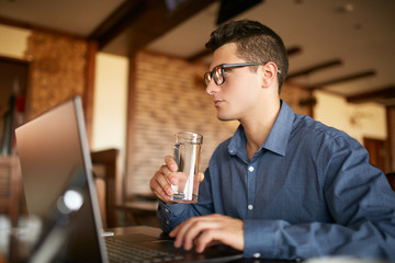 Handsome caucasian man with glass of water in hand works with laptop. Businessman in glasses drinks water for body hydration while working. Attractive designer quench thirst. Healthy lifestyle theme.