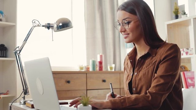 Young graduate of prestigious university writing thesis at home. Beautiful smiling girl in glasses sitting at the laptop, working. Daytime.