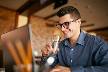 Smiling caucasian man with glass of water in hand works with laptop. Businessman in glasses drinks...