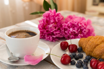 Close up of morning coffee, croissant, blueberries and beautiful pink peony flowers on light table. Cozy breakfast on Mother or Woman day.