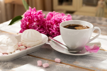 Close-up of morning coffee, marshmallows and beautiful pink peony flowers on light table. Cozy breakfast on Mother or Woman day.