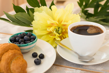 Close-up of morning coffee, croissant, blueberries and beautiful yellow peony flowers on light table. Cozy breakfast on Mother or Woman day.