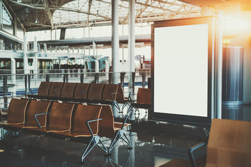 Blank white billboard placeholder mockup in shopping mall; empty informational banner in airport waiting hall or waiting room of railway station depot surrounded by rows of empty wooden seats