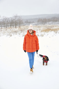 Full length shot of a happy woman walking her small dog outdoors on snowy landscape in winter.