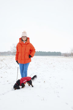 Full length shot of a happy woman walking her small dog outdoors on snowy landscape in winter.