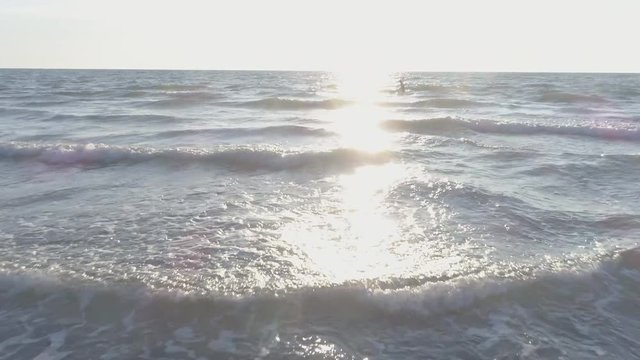 Young man surfer wearing wetsuit carrying surf board walking in the calm ocean water at sunset aerial drone dolly