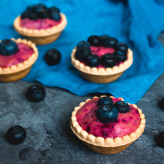 Sweet mini tartlets with cream and berries on a blue linen cloth. Simple dessert snack recipe with blueberry. Summer food concept.