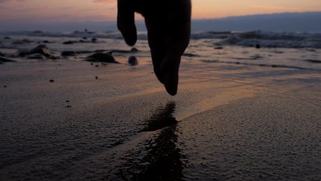 Beautiful sunset on the sea coast. On the sand, he draws a heart shape with his finger. The camera is very close to water and sand. The focus is at the very nearest point.