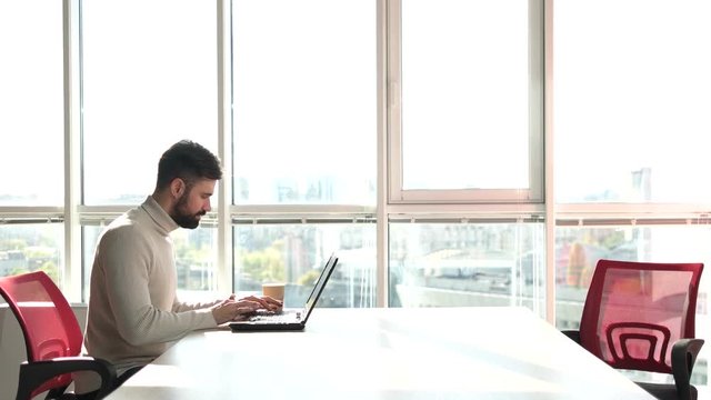 Young bearded man working as freelancer on laptop. Man working on laptop in office with windows background.