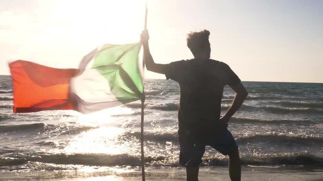 Young man in swimming trunks waving italian flag on a pole on the sea shore at the beach at sunset silhouette